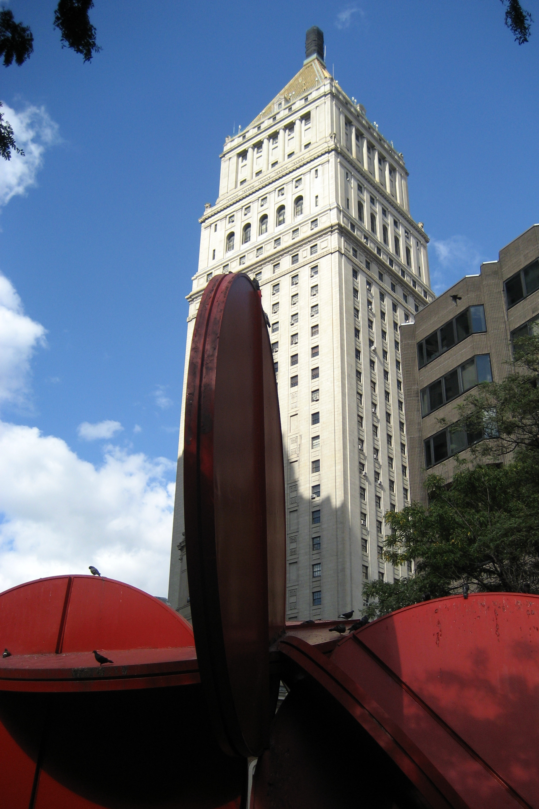 NYC: Police Plaza, Five in One and US Courthouse The NYPD is headquartered at One Police Plaza (1PP), located on Park Row across the street from City Hall and through the Municipal Building’s archway. Prominently centered in the plaza is Bernard Rosenthal’s Five in One (1974), standing at 30 feet high and made of 75 tons of Cor-ten steel. It is now painted bright red, according to Rosenthal’s original design (although lack of funds left the statue unpainted for years after it was completed). Rosenthal calls the sculpture abstract, but many people choose to see the five boroughs intertwined symbolically. The U.S. Courthouse was built as the Federal courthouse in New York City. Design work was started in 1933 and, after Cass Gilbert's (Woolworth Building) death the next year, supervised by his son until its completion in 1936. The six-storey base of the building is reminiscent of a Classical temple with its pilastered facades and colonnaded entrance, and rising from this is the sturdy 32-storey tower, topped by a pyramidal roof clad in gilded terra-cotta and a lantern.