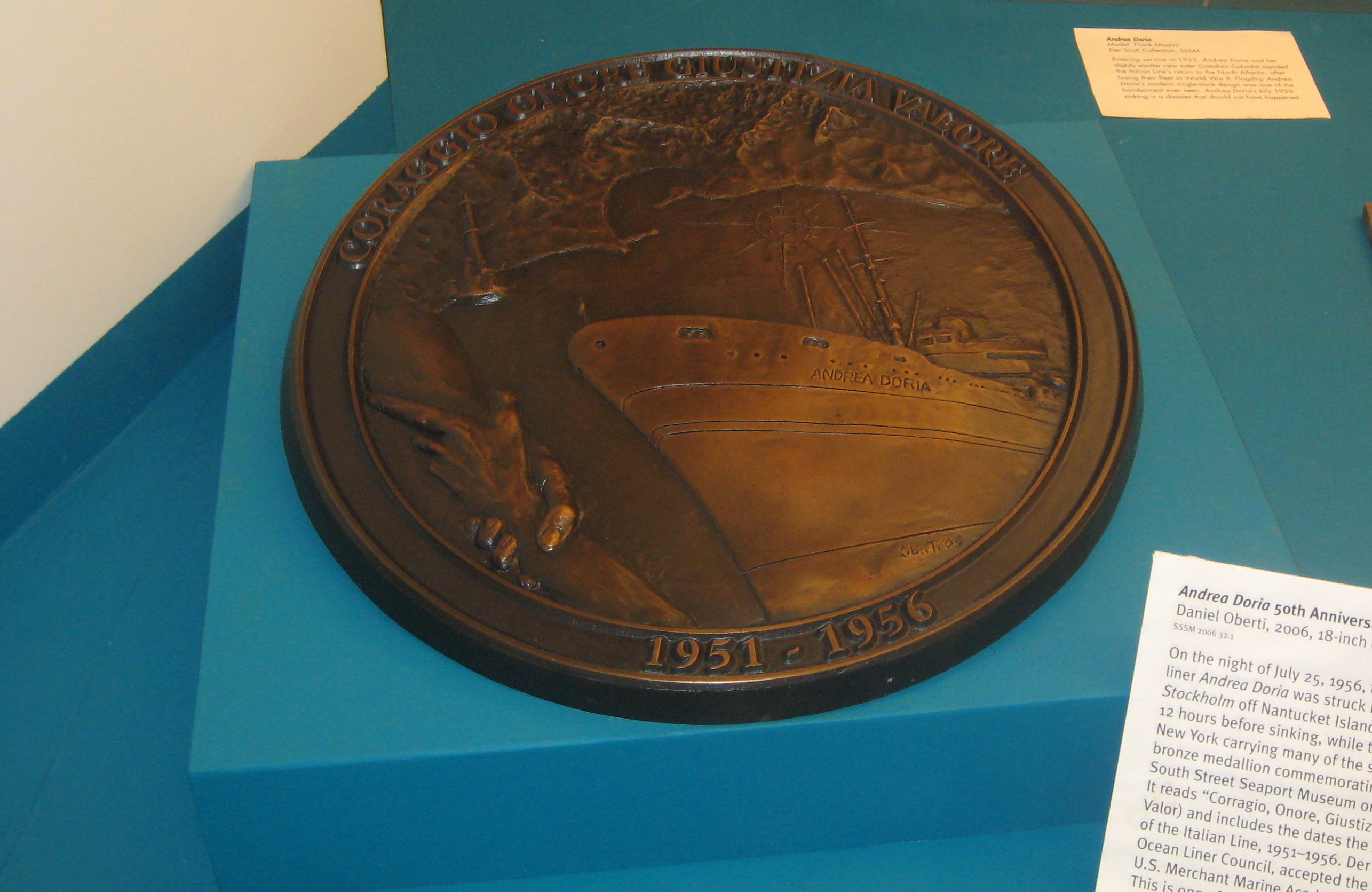 NYC - South Street Seaport Museum - Andrea Doria 50th Anniversary Medallion Andrea Doria 50th Anniversary Medallion Daniel Oberti, 2006, 18-unch diameter, bronze SSSM 2006 32.1 On the night of July 25, 1956, the New York-bound Italian ocean liner Andrea Doria was struck by the Europe-bound Swedish liner Stockholm off Nantucket Island. The Doria remained afloat for 12 hours before sinking, while the Stockholm was able to return to New York carrying many of the survivors of the Italian ship. This bronze medallion commemorating the tragedy was presented to South Street Seaport Museum on the 50th anniversary of the sinking. It reads "Corragio, Onore, Giustizia, Valore" (Courage, Honor, Justice, Valor) and includes the dates the Andrea Doria served as the flagship of the Italian Line, 1951-1956. Der Scutt, Chairman of the Museum's Ocean Liner Council, accepted the medallion at a ceremony held at the U.S. Merchant Marine Academy in Kings Point, NY on July 23, 2006. This is one of two identical medallions; the second is in the collection of the Galata Museo del Mare in Genoa, Italy. The medallions were sponsored by Jerome Reinert, Angela E. Addario and Pierette Simpson.