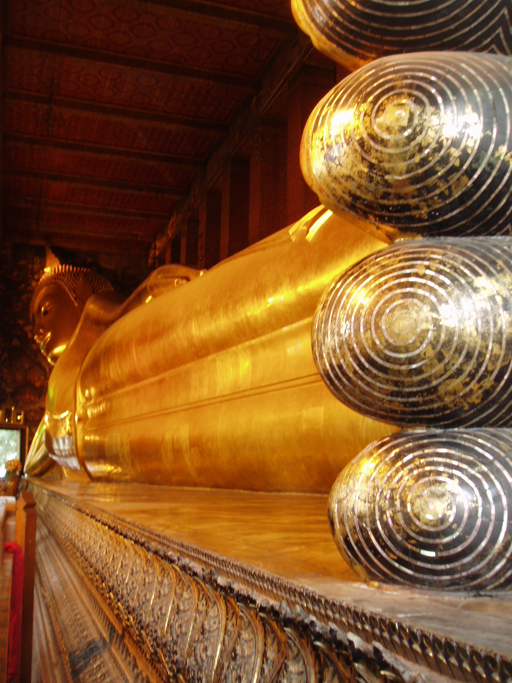 Reclining Buddha, Wat Pho, Bangkok Wat Pho is the largest and oldest temple complex in Bangkok, and along with the temmple of the royal palace the most visited. The sprawling complex covers a huge area and it is quite easy to lose oneself within it's series of courtyards, gateways and stunning temple buildings. The present complex largely dates back to 1788, having been founded in response to the sacking of the former capital Ayutthaya by the Burmese in 1767 where a similar complex was destroyed. It is known as a place of learning and has a renowned school of Thai massage within the site. The temple complex is entered through one of many gateways guarded by huge Chinese statues, believed to have been imported as ballast on ships trading with China..The outer gates have fierce warrior figures, whilst the inner courtyards have the bizarre 'farang' ('foreigner') figures with their peculiar 'top hats', believed to represent the first European visitors to the east. Many of the inner courtyards are surrounded by a cloister containing over 400 sculptures of the seated Buddha, whilst the main Bot, the spiritual nucleus of the temple, is beautifully decorated within with frescoes of rich landscapes in red and gold. The biggest and most spectacular attraction of the temple however is the Hall of the Reclining Buddha, housing an enormous gilded recumbent figure of the Buddha over 160ft long, filling the entire centre of the chamber. en.wikipedia.org/wiki/Wat_Pho