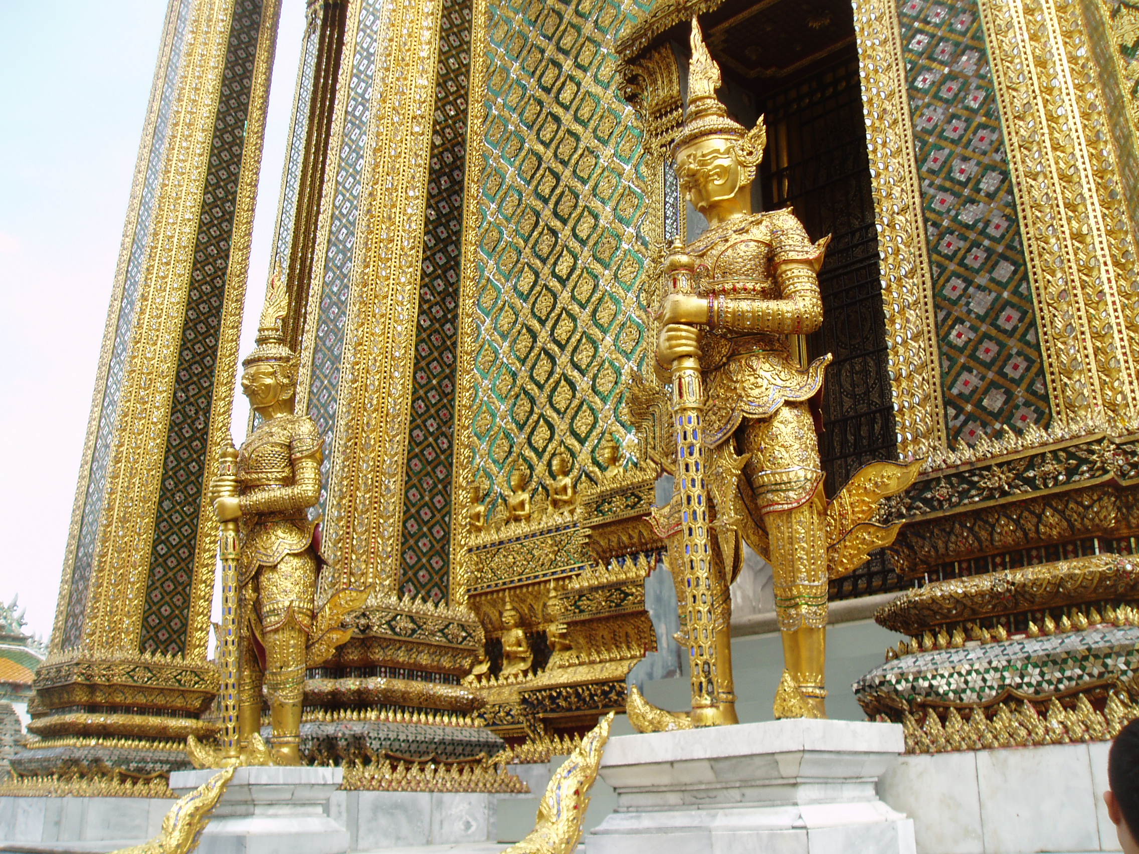 Golden Guardians, Wat Phra Kaew, Bangkok Bangkok's most unmissable attraction is of course the Grand Palace, and most specifically the temple complex of Wat Phra Kaew (Temple of the Emerald Buddha), famed for it's riot of coloured and gilded ornament, paintings and sculptures; quite simply, so visually stunning there is nothing quite like it anywhere else! The Palace and Temple complex were begun in 1782, the year the capital was moved to Bangkok, and parts of the palace buildings betray 18th century European influence combined with traditional Thai style, such as the breathtaking gilt spires on the roof. Most of the interiors of the Palace itself are off limits to visitors since, although no longer the main residence of the thai monarchy, it is frequently used for state functions and ceremonies. The Wat Phra Kaew complex however is the greatest draw, famed for it's stunning architecture and the famous 'Yaksha' guardian figures that flank all the main entrances to the complex. These towering figures, with their rich colours and tapering crowns, represent demonic characters from the mythological epic the 'Ramakien', and are identifiable as distinct individuals, all here serving a benign, protective role. The Ramakien is also the subject for a stunning sequence of wall paintings within the cloister that encirlces the entire site, illustrating in minute detail the battles of the heroic monkey warriors, led by the monkey god Hanuman, against the demonic armies and kingdoms of Tosakan. The Temple of the Emerald Buddha itself forms the largest structure and contains the venerated (though small) Buddha image. The complex contains several other iconic buildings clad in sumptuous decor, most notably the library or 'mondop' with it's gilt spire along with the great golden stupa. The temple complex is technically a royal chapel rather than a working monastery like most Thai temples as it has no resident monks (the sheer volume of visitors leaves little room for anyone else anyway!). en.wikipedia.org/wiki/Wat_Phra_Kaew en.wikipedia.org/wiki/Grand_Palace