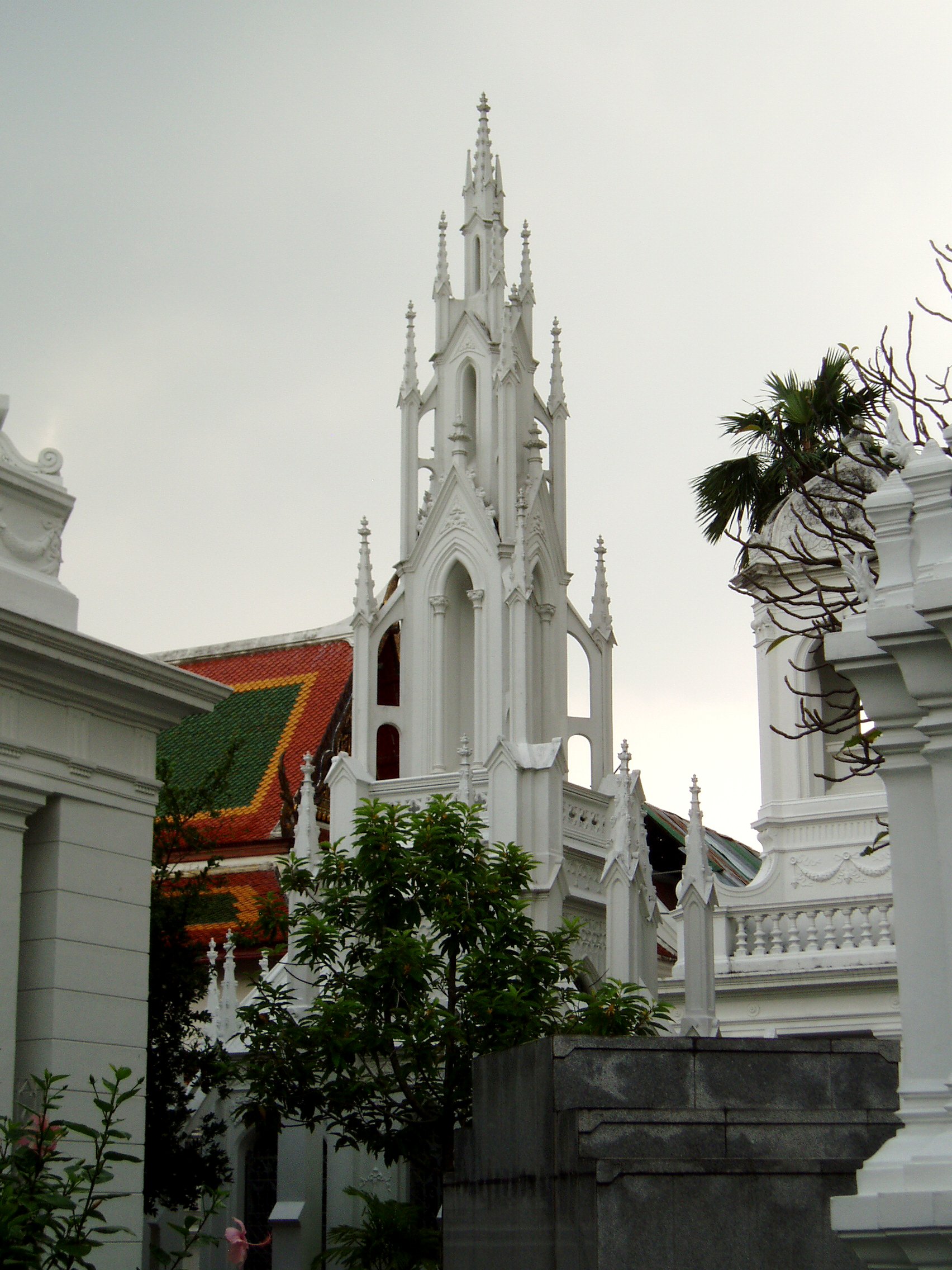 Gothic Steeple, Royal Cemetery, Wat Ratchabophit, Bangkok Wat Ratchabophit is one of the most unusual temples in Bangkok; built in the latter half of the 19th century it shows marked European influence in certain areas, such as the neo-gothic interior of the main bot (prayer hall) and many of the monuments in the adjoining Royal Cemetery, that includes a domed classical style tower and pinnacled gothic steeples amongst more traditional Thai and Khmer style structures. The centrepiece of the Temple is the huge tile-covered stupa that grows from the heart of the complex and is surrounded by a tight circular courtyard. The surrounding structures are richly decorated and gilded with typical Thai richness. The entrance gates to the temple compound have carved and painted door panels with bizarre figures that appear to be 'farang' (foreign/European) guards holding rifles! en.wikipedia.org/wiki/Wat_Ratchabophit