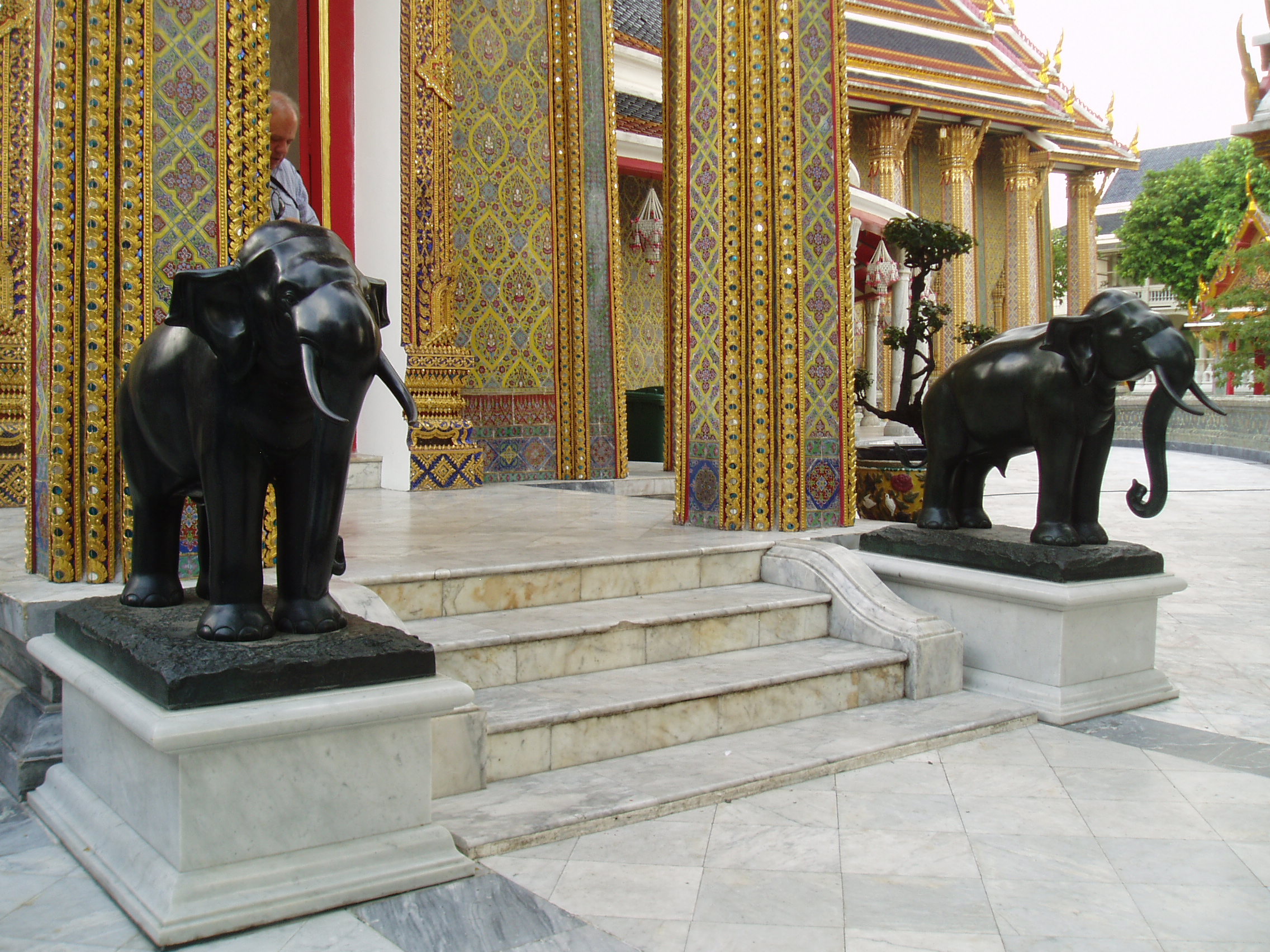 Elephants, Wat Ratchabophit, Bangkok Wat Ratchabophit is one of the most unusual temples in Bangkok; built in the latter half of the 19th century it shows marked European influence in certain areas, such as the neo-gothic interior of the main bot (prayer hall) and many of the monuments in the adjoining Royal Cemetery, that includes a domed classical style tower and pinnacled gothic steeples amongst more traditional Thai and Khmer style structures. The centrepiece of the Temple is the huge tile-covered stupa that grows from the heart of the complex and is surrounded by a tight circular courtyard. The surrounding structures are richly decorated and gilded with typical Thai richness. The entrance gates to the temple compound have carved and painted door panels with bizarre figures that appear to be 'farang' (foreign/European) guards holding rifles! en.wikipedia.org/wiki/Wat_Ratchabophit