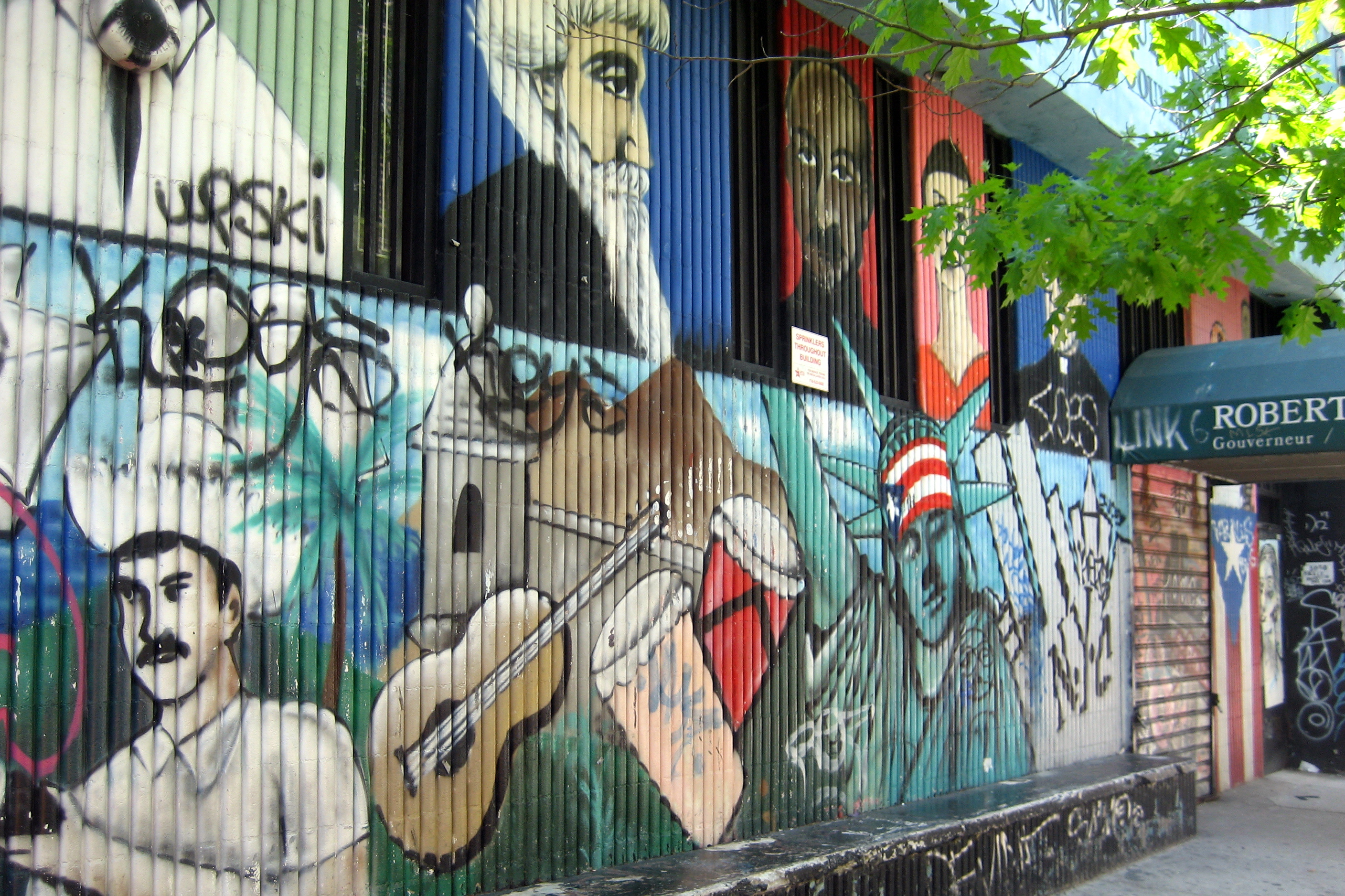 NYC - East Village: Roberto Clemente Center - mural The mural outside the Roberto Clemente Center was painted in 1996 by local artist Antonio Garcia, known simply as Chico. The mural represents the mixing of Latin American cultures, races, and identities up to the current Period of immigration to North America. The mural is done on two levels. The lower levels depcicts symbols of the evolution of cultures. On the left (off camera) it represents Native Americans followed by the arrival of the Spanish. The mixing of those two races resulted in the evolution of the peasant, Jibarao (depicted with hat and machete). The Jibaro's culture is represented by string musical instruments. Similarly, the African influence in Latin nationality is represented by the drums. These cultures and races mixed and resulted in the racially mixed "trigueno" nation. Following that is the representation of immigration to the U.S. in search of economic and other freedomds--Statue of Liberty. The top lefvel of the mural depicts eight outsanding symbols of a racially integrated Lain American pride: men and women; "triguenos," black and white; Dominican, Puerto Rican, Mexican, Venezuelan, Cuban, Salvadorean--all representatives of the honor of being South and Central American, Mexican and Carribean. From left to right are (not pictured): Dominican Indian Leader Anacona; Mexican Constitutional Reformer Benito Juarez, Liberator of South America Simon Bolivar, Puerto Rican feminist Luisa Capetillo, and (pictured) Puerto Rican liberation leader Ramon E. Betances, Cuban independence leader Jose Marti, Puerto Rican poet Julia de Burgos, Salvadorean leader of the rights of the poor Archbishop Romero. To the extreme right, on the other side of the door, is the namesake, baseball Hall of Famer, Roberto Clemente. The Roberto Clemente Center was first established in 1982 as an outpatient mental health service: It was the first mental health program in New York State to propose that culture is an essential component of mental health treatment. Cultural sensitivity was operationalized by including an all bilingual and bicultural staff. The importance of culturally sensitive care is now well established in the mental health field. The Clemente Center broke ground in making this possible for other mental health groups. The Roberto Clemente Center was made possible by the advocacy efforts of the Minority Advisory Council of the New York State Office of Mental Health and especially by the energetic support received from community-based organizations and leaders. The Center was first funded by the New York State Office of Mental Health. The Center was named after Roberto Clemente in order to honor the values and work of the late Pittsburgh Pirates baseball star. Clemente spoke out about the discrimination that African-American and Latino players suffered in baseball. Throughout his life, Roberto Clemente strove for the highest performance goals and expectations. His love for children led him to develop children's sports programs. Clemente is also known for his humanitarian and relief efforts. Roberto Clemente died in a tragic plane crash on New Year's Eve of 1972 while transporting medical relief for the victims of an earthquake in Nicaragua. The neighorhood is filled with Chico's renegade work which dates back to the early 80's when he used to tag old redbird subway cars. He is often credited with transforming subway graffiti into a more accepted art form. His first mural — long gone — was a jab at then President Reagan: a tank driving toward the words "World War III." Then local businesses offered to pay him $100, then more, to paint walls near their stores creating an eclectic range of art.