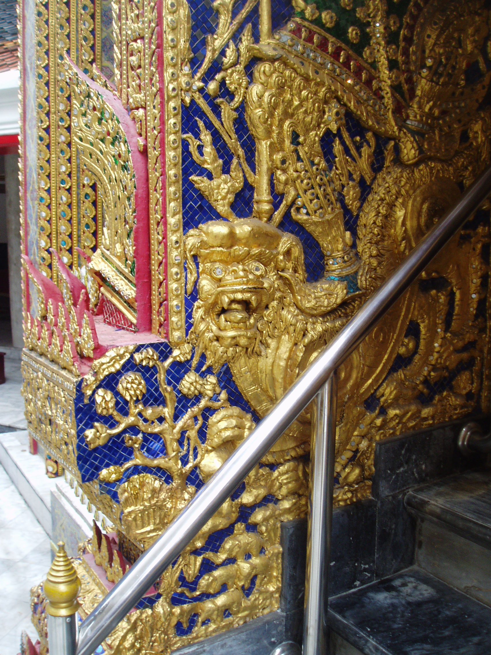 Gilded Relief, Wat Ratchabophit, Bangkok Wat Ratchabophit is one of the most unusual temples in Bangkok; built in the latter half of the 19th century it shows marked European influence in certain areas, such as the neo-gothic interior of the main bot (prayer hall) and many of the monuments in the adjoining Royal Cemetery, that includes a domed classical style tower and pinnacled gothic steeples amongst more traditional Thai and Khmer style structures. The centrepiece of the Temple is the huge tile-covered stupa that grows from the heart of the complex and is surrounded by a tight circular courtyard. The surrounding structures are richly decorated and gilded with typical Thai richness. The entrance gates to the temple compound have carved and painted door panels with bizarre figures that appear to be 'farang' (foreign/European) guards holding rifles! en.wikipedia.org/wiki/Wat_Ratchabophit
