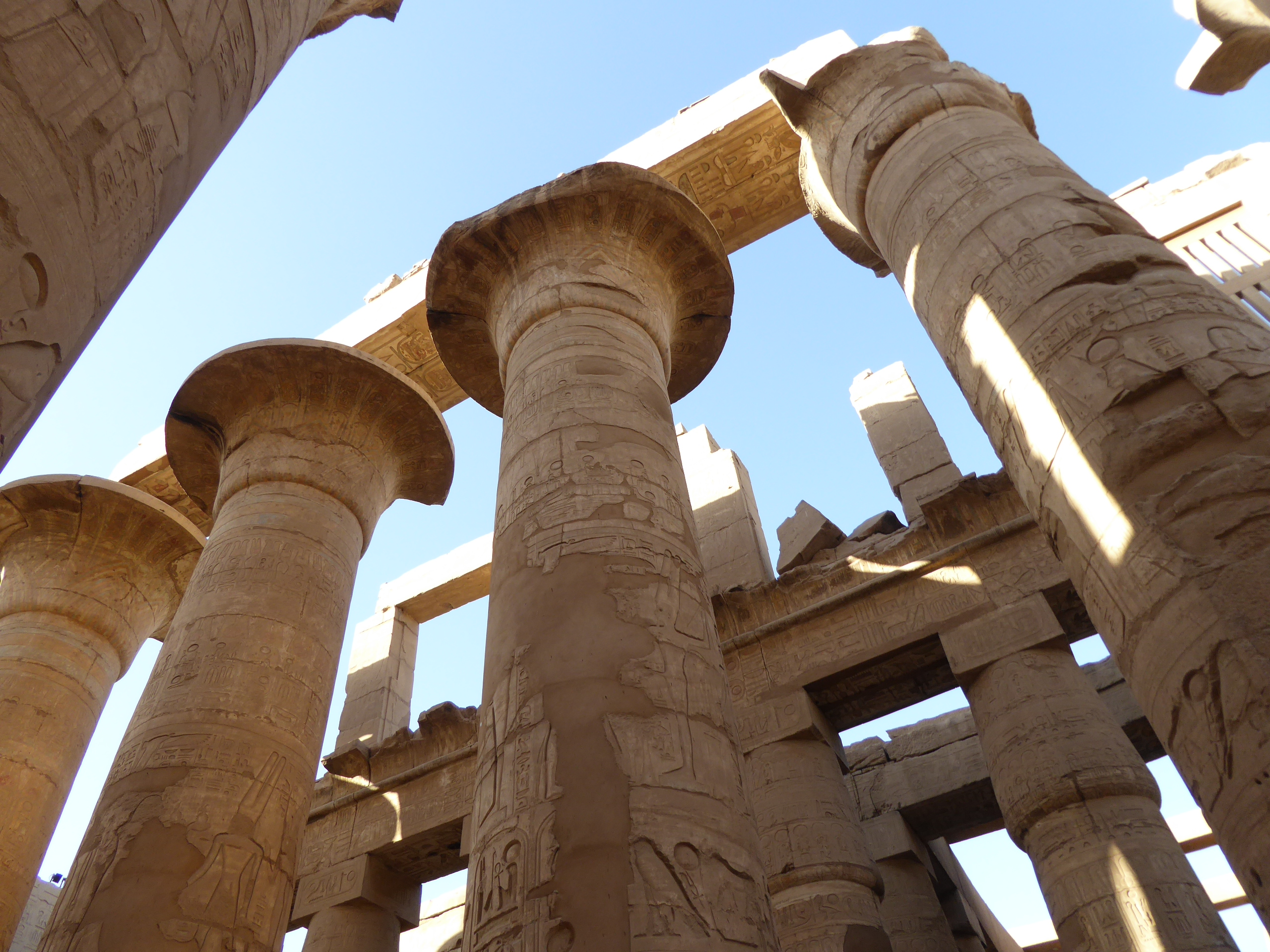 Hypostyle Hall, Karnak One of the grandest and most rewarding monuments of the ancient world, the Temple of Amun at Karnak simply cannot fail to impress with its grandeur. It is a vast complex of buildings over three main precincts, the greatest of which is that of Amun with the enormous great temple at its heart. This is the main draw for all visitors to Karnak, with its avenue of sphinxes, huge courtyards and incomparable pillared hypostyle hall. It is not only one of Egypt's most important monuments but simply one of the greatest ancient sites anywhere. The great temple of Amun dominates the complex and is the work of successive dynasties throughout the New Kingdom, at which point the local god Amun had been established as the state god of Egypt (his name means 'the hidden one', as he only rose to prominence relatively late in Egyptian history, largely connected to the rising importance of Thebes as the centre of power, thus its local god rose with it). Some of the earliest parts at the east end date to the reign of Thutmoses I with successive sections added by other 18th & 19th Dynasty rulers, with great obelisks erected by Hatshepsut and much of the Hypostyle Hall and pylons built under Seti I and his son Ramesses II. The first courtyard and the main pylon were the latest additions to the temple under the reign of Nectanebo I and were never fully finished. The temple is approached through and avenue of large ram-headed sphinxes, each protecting a small pharoah figure between their paws, representing the god Amun (whose sacred animal was the ram) protecting the king. These statues are usually identified as dating to the reign of Ramesses II, but it is possible they may date further back to Amenophis III. Originally this avenue stretched all the way through what is now the first courtyard to the 2nd pylon and hypostyle hall, but after construction of the 1st pylon and courtyard those beyond the new facade were stored in rows either side of the new courtyard beyond the entrance pylon. The main facade is formed by the towering 1st pylon built under Nectanebo I and never finished, the stonework is still rough-hewn and lacking in decoration and the northern tower remains somewhat shorter than its southern neighbour. The southerrn tower has at its rear the remains of a mud-brick ramp used during construction. The courtyard beyond is vast and dotted with pillars and statues from various earlier stages in the growth of Karnak, including a towering column from a pavilion built by Nubian Pharoah Taharqa, two colossi of Ramesses II (one usurped later by Pinedjem) and more of the ram-sphinxes that once lined the approach. Beyond the courtyard is the more ruinous 2nd pylon which leads to the most famous part of the Karnak Temple complex, the incredible Hypostyle Hall, sometimes called the 'Hall of a Hundred Columns' owing to the great multitude of thick, round columns in every direction, like a forest of enormous tree-trunks in stone. There are 134 columns in all in 16 rows, the central axis columns being taller with palm-capitals, whilst the rows either side of this carried a clerestorey of stone-latticed windows, many of which survive (originally these would have been the main light source when the hall had its roof). The decoration on the columns and side walls dates mostly to the reign of Ramesses II and is in sunken relief, but some earlier work from Seti I's reign in raised-relief can be seen on the north side. Continuing along the main eastern axis the great temple becomes more ruinous and harder to follow, but several major features stand out such as the obelisk of Hatshepsut and the much later barque shrine, whilst some distance beyond the festival hall of Thutmosis III, a pillared structure that represents the most substantial remaining part of the eastern sections of the temple and retains some coloured decoration inside. In addition to the main eastern axis of the temple there is the southern axis that is formed of four courtyards and further pylon towers and gates which extend towards the precinct of Amun's consort Mut (a much more ruined site reached by equally ruined sphinx avenues to the south, one of which also connects with that leading to Luxor Temple a mile or so further on).. These sections of the complex are mostly closed to visitors at present with stabilisation works ongoing. There are further smaller temples and areas of great interest around the huge Karnak site, which we have visited on a previous trip (we once spent an entire day here in the pre-digital 1990s), but sadly time was limited on this occasion to focusing on the major sections of the great temple itself, so we hope to return to spend longer here another time. en.wikipedia.org/wiki/Karnak